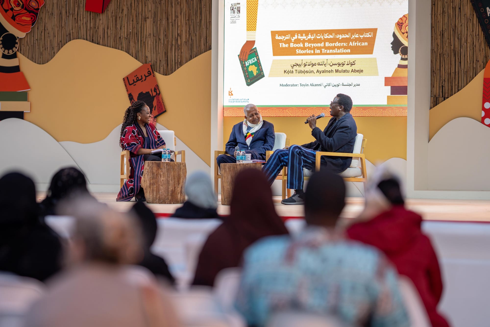 Photo taken by audience member of a dais decorated with African themes, three people on a panel, behind them a sign in Arabic and English, reading "The Book Beyond Borders: African Stories in Translation: Kọ́lá Túbọ̀sún, Ayalneh Mulatu Abeje, Moderator: Toyin Akanni