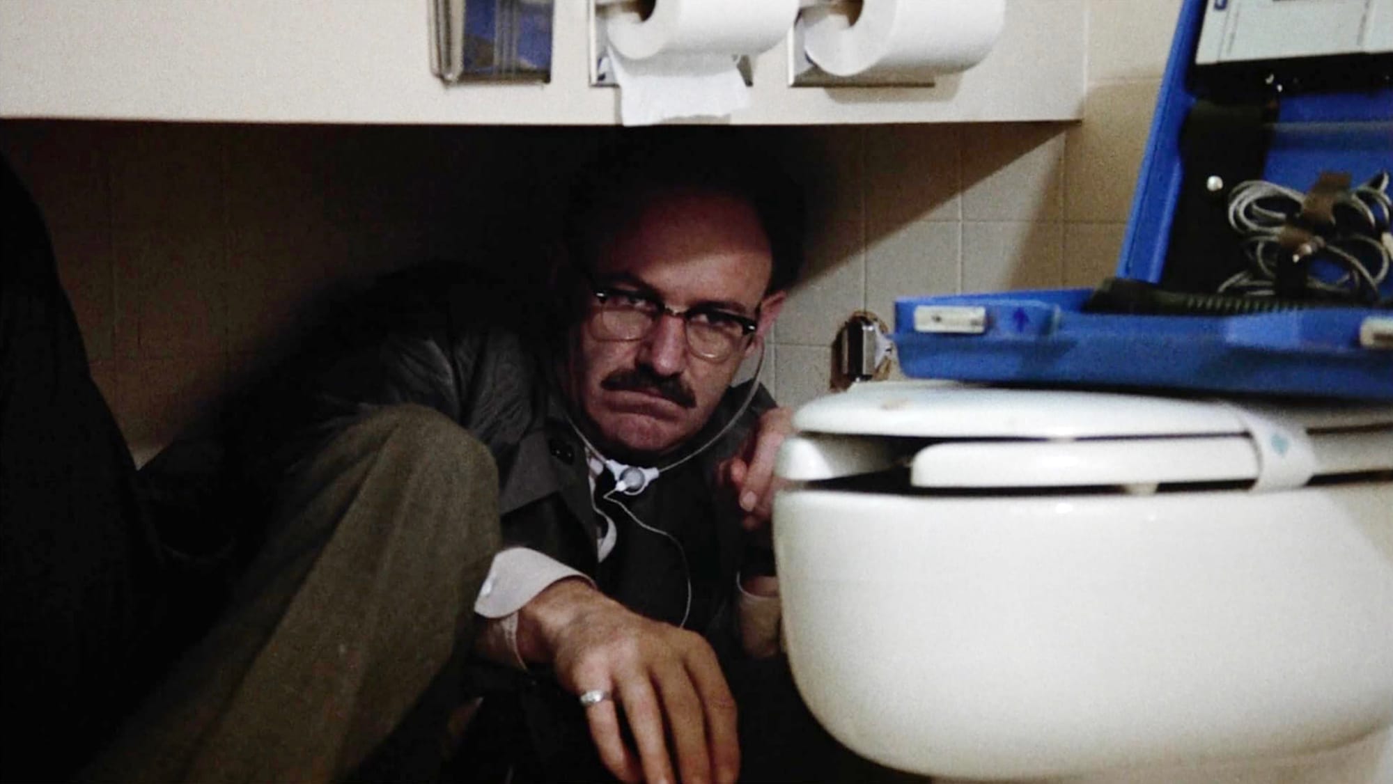 Hackman's character "Harry Caul" using audio surveillance equipment in a bathroom and he is lying down under the toilet paper rack next to the shitter tuning his listening device.