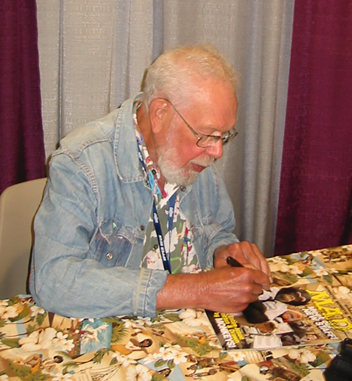 ALT: An older Jaffee seated at a table, wearing specs, a convention lanyard, and a jeans jacket over a colorful shirt, signs the cover of an issue of MAD