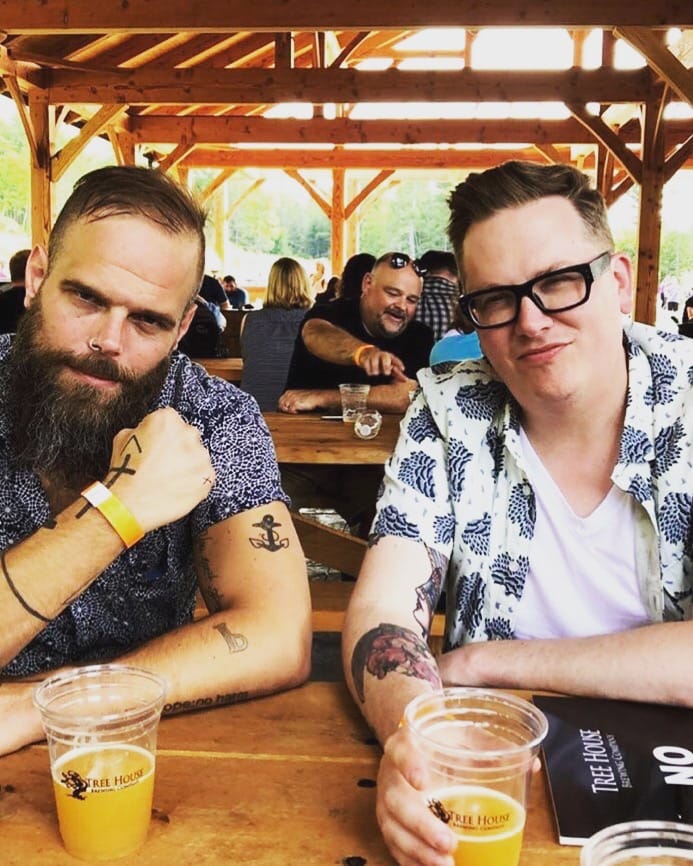 Two men with tattooed forearms in blue-and-white printed short-sleeved button-front shirts at a Tree House brewery, seated outdoors at a long wooden table, drinking fancy beer