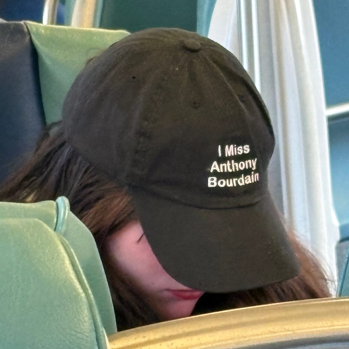 Person with head tilted down so you can't see their face. They are wearing ballcap reading "I Miss Anthony Bourdain"