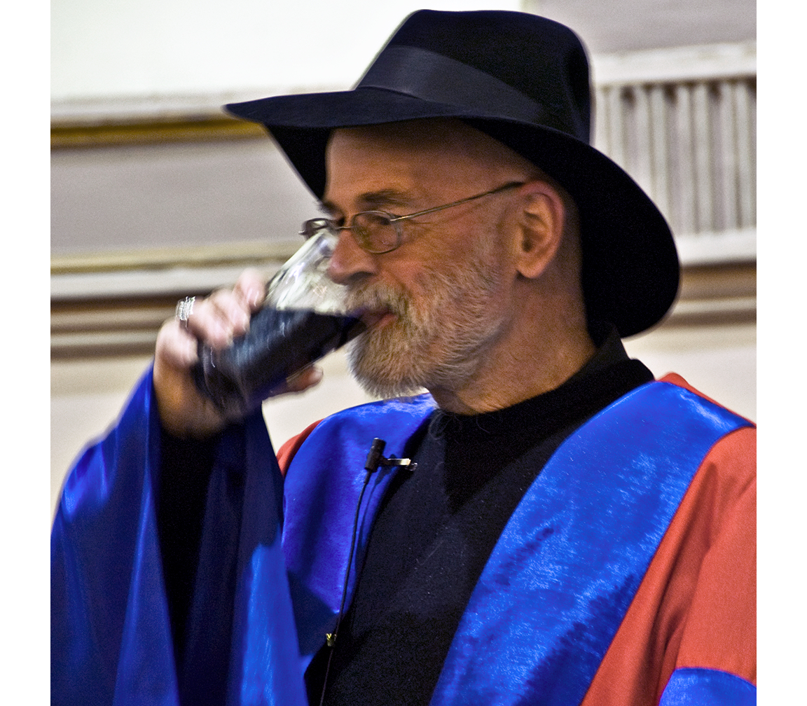 Pratchett in bright red and blue robe and a wide-brimmed black hat happily guzzling a pint.