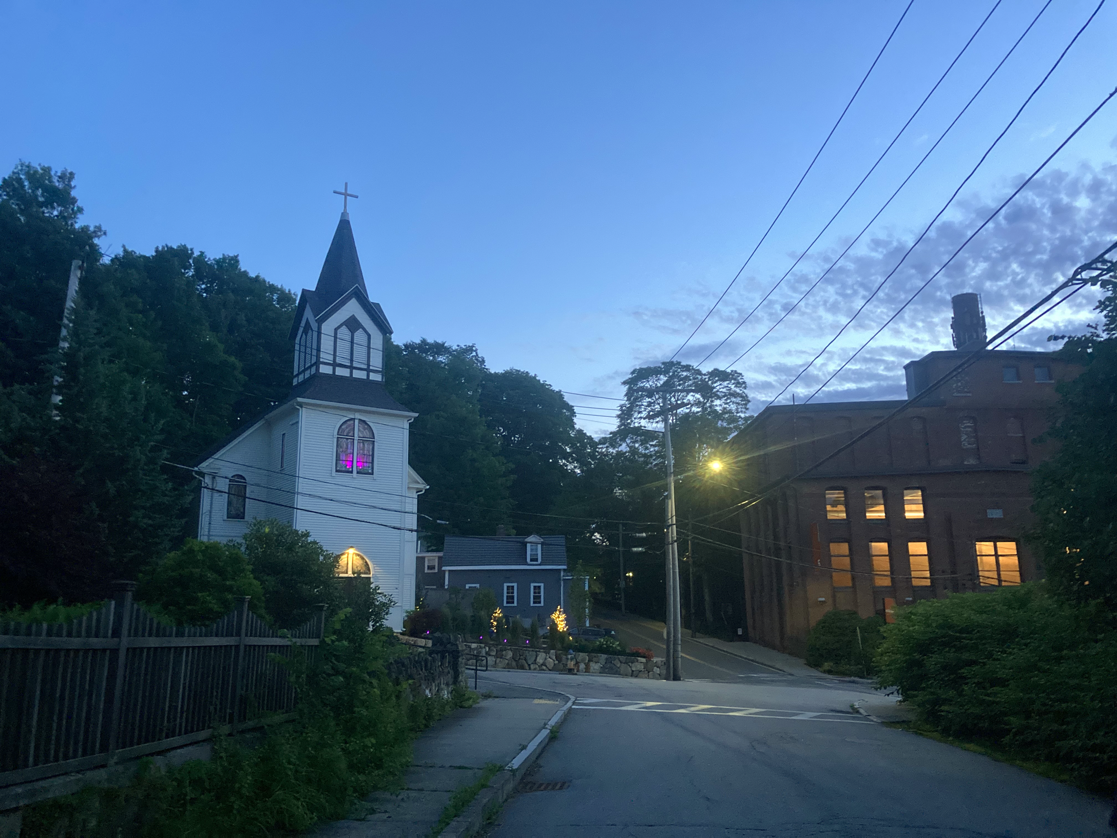 An old church with spooky pink lighting upstairs at twilight with dappled, darkening clouds; telephone poles and wires, a lit streetlamp