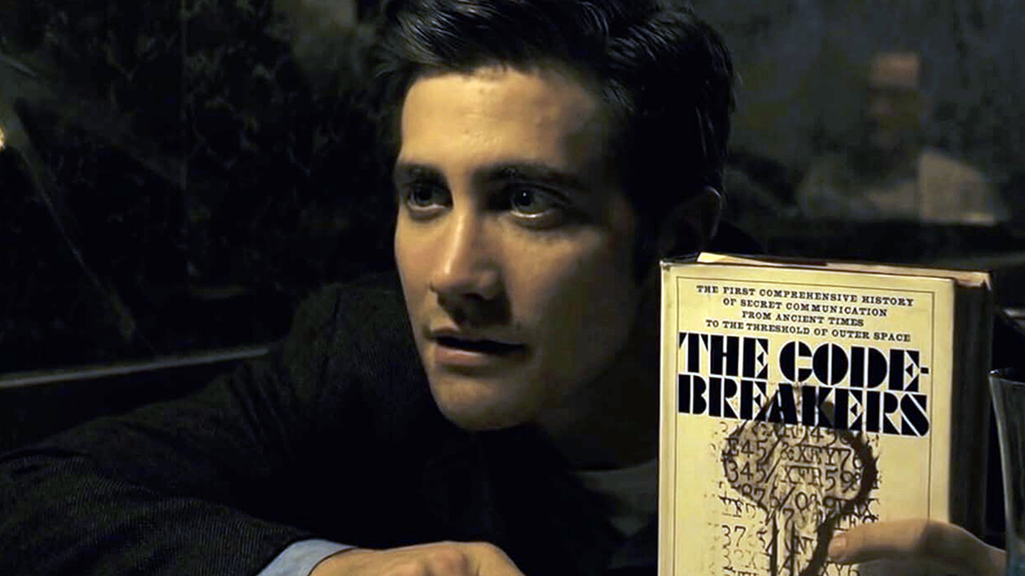 Wide-eyed Eagle-scout-looking Jake's character holding a copy of "THE CODE BREAKERS: THE FIRST COMPREHENSIVE HISTORY OF SECRET COMMUNICATION FROM ANCIENT TIMES TO THE THRESHOLD OF OUTER SPACE"