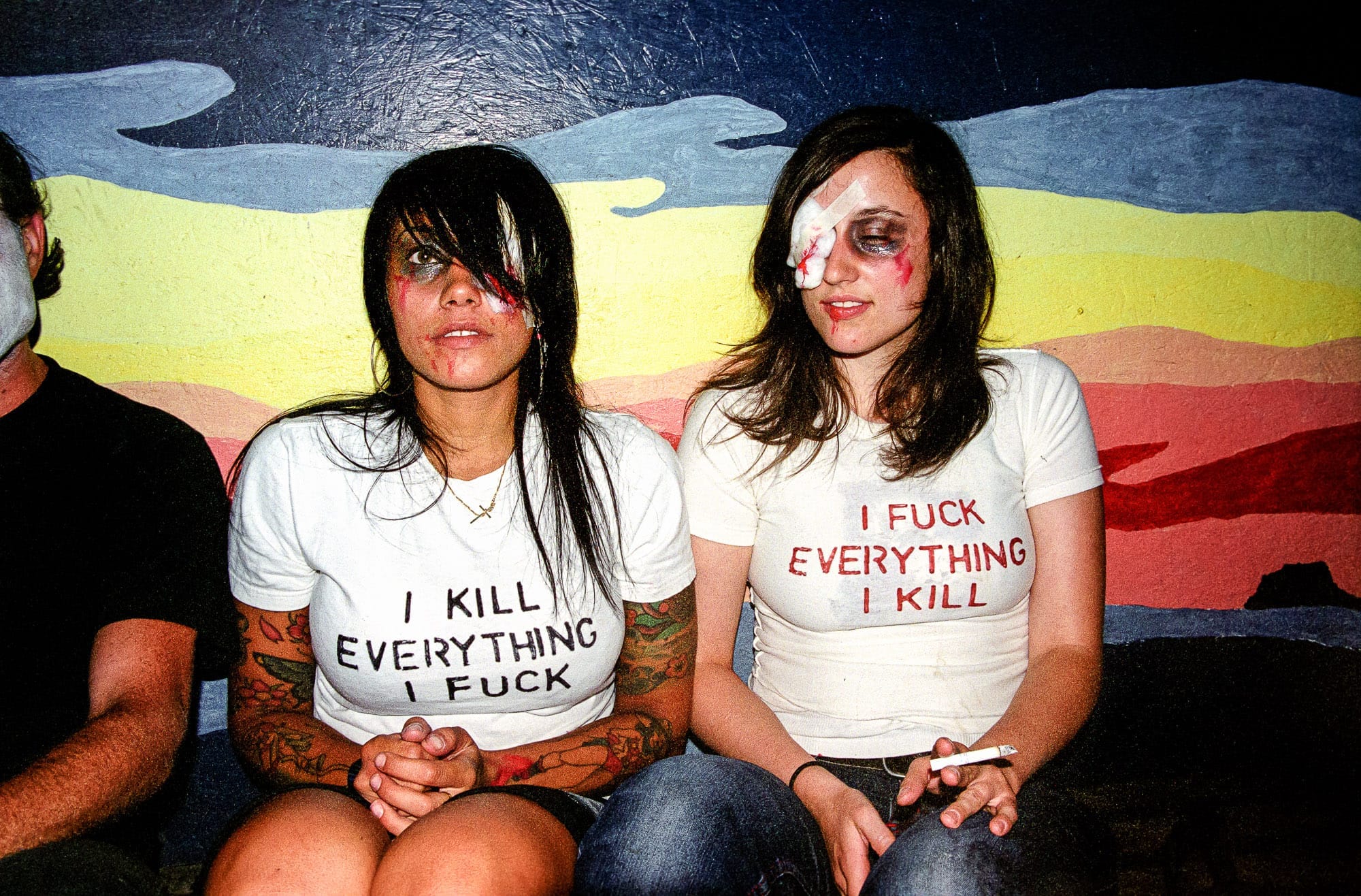 Two long-haired girls in horror makeup (black eyes, bandages, fake blood) in their late teens or early 20s, at a party, wearing white t-shirts reading in block letters, I KILL EVERYTHING I FUCK and I FUCK EVERYTHING I KILL. They are seated against a colorful wall painted with an abstract landscape. One girl is holding a lit cigarette