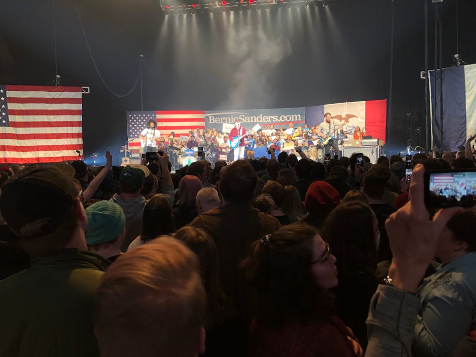 Ezra Koenig strums guitar on large stage. Crowd of 50+ people sit on risers behind, more facing him. Above Ezra there's American Flag, Iowa state flag, and sign reads: BernieSanders.com