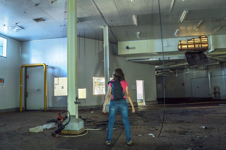 A young person in t-shirt and jeans, carrying a backpack, stands in reverie before the decaying remains of a factory