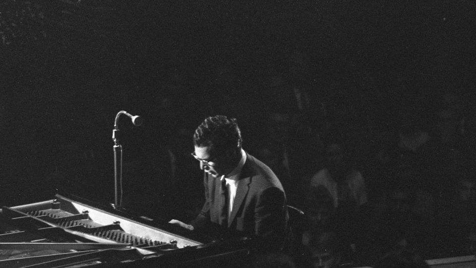 Black and white image of Dave Brubeck seated at the piano in near-darkness, in concert in 1959 