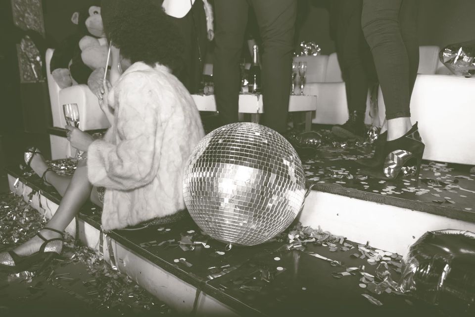 A lady in black satin heels and a white plush jacket seated next to a mirror ball on the steps of a nightclub