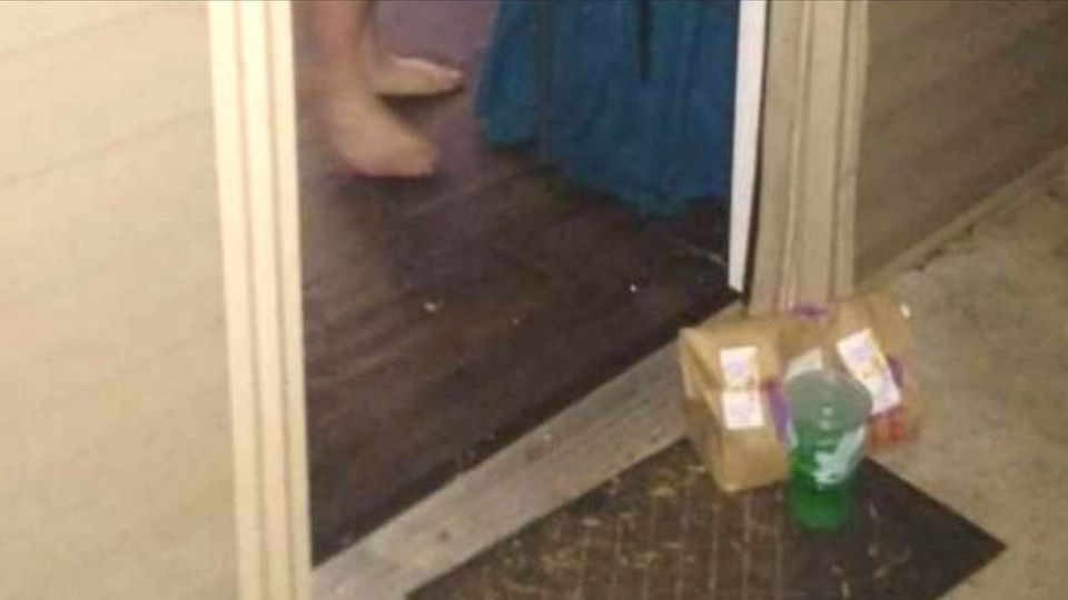 A doorstep, the door open, and bare feet visible. On the doormat, a paper delivery bag and a large plastic drink cup 