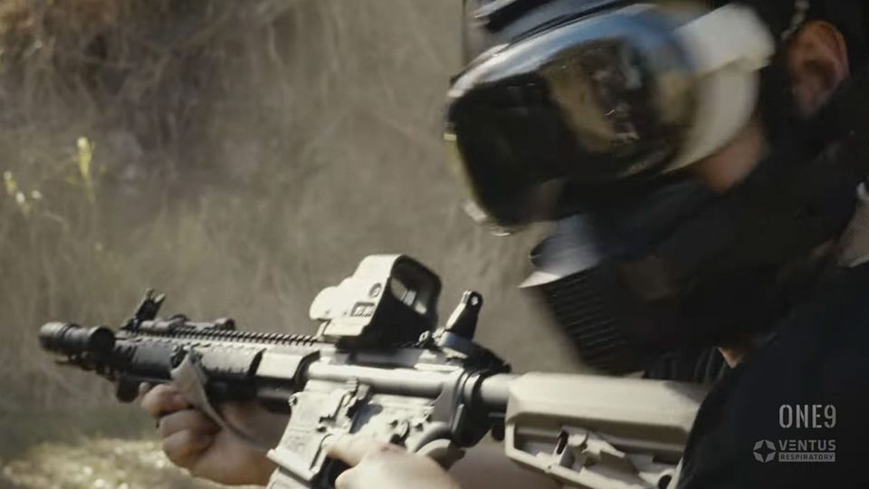 A man wearing an Apple Vision Pro, together with a military helmet and respirator; he is holding an automatic weapon