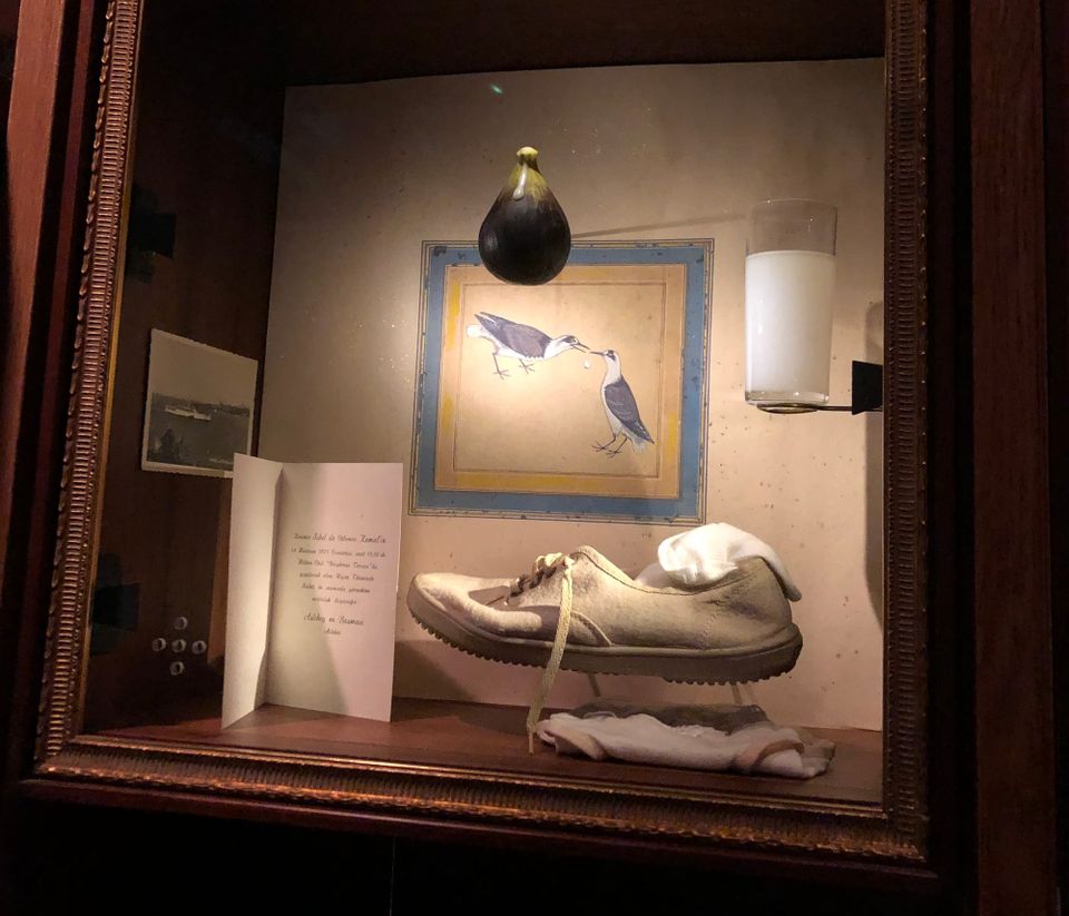 a shadow box holds a single worn white sneaker, a holiday card, glass of cloudy white raki, and a fig hanging from the top.