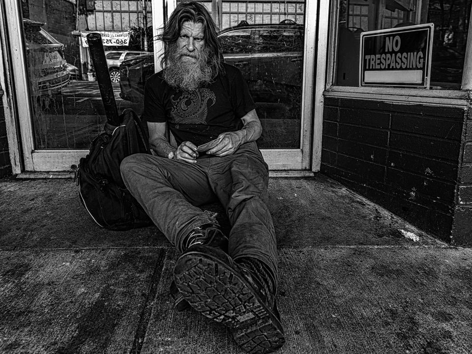 "Alone," a black and white photo of an older, hard-bitten, unshaven man seated in a doorway on the street