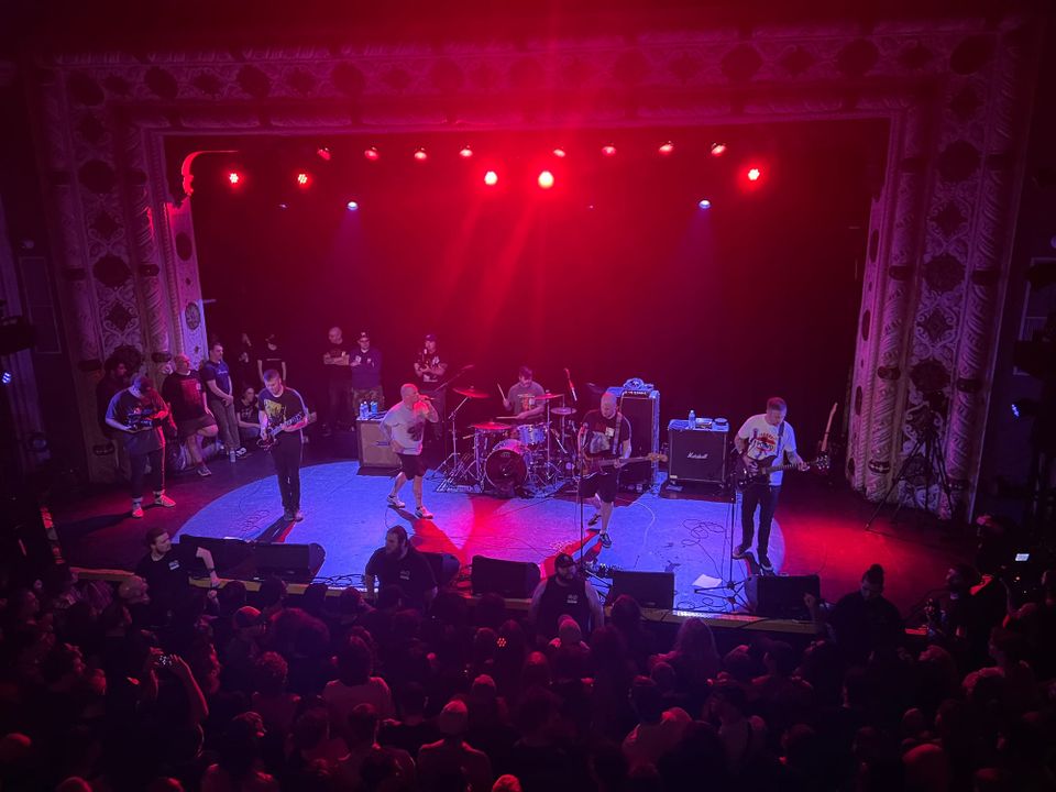 A punk band onstage under lurid red lights, playing to a packed crowd in a big venue