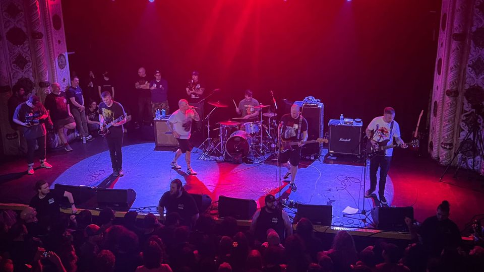 A band performing on a darkened under ruby-red lights before a packed crowd