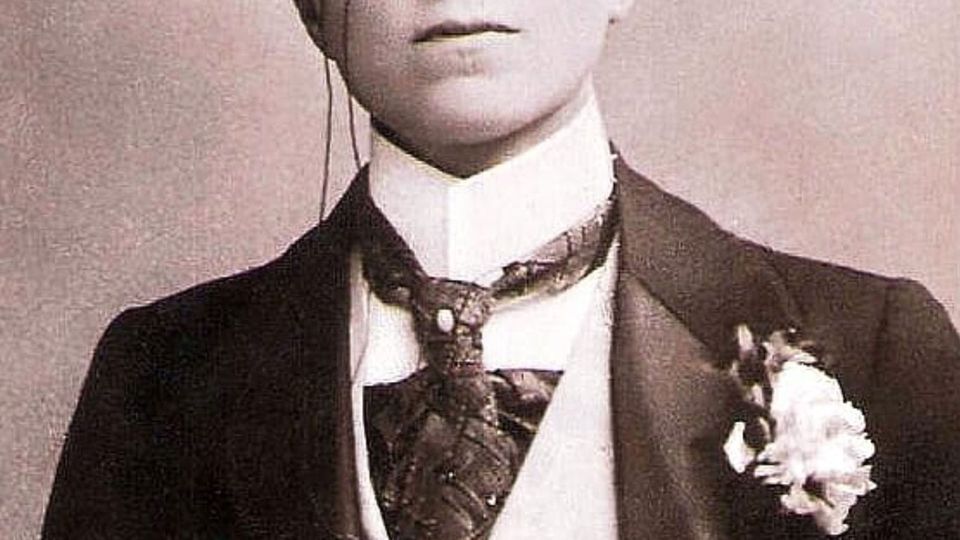 The lower face of a woman in an early 20th-century man's suit with cravat, tie pin and boutonnière 