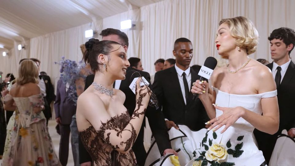 Emma Chamberlain, in a brown lace gown, tilts her head and smiles at Gigi Hadid, in a white ballgown