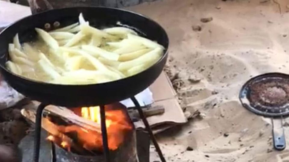 Sliced potatoes boiling in a small iron pan over a wood fire on a wire brazier in the sand