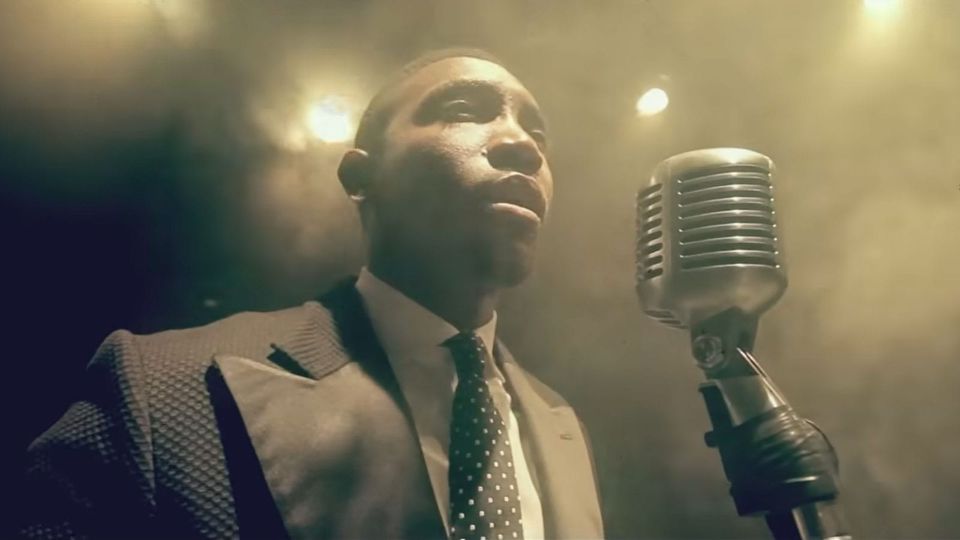 Timi Dakolo performing "Great Nation" in a suit with wide lapels and polkadot tie, before a big old condenser mic