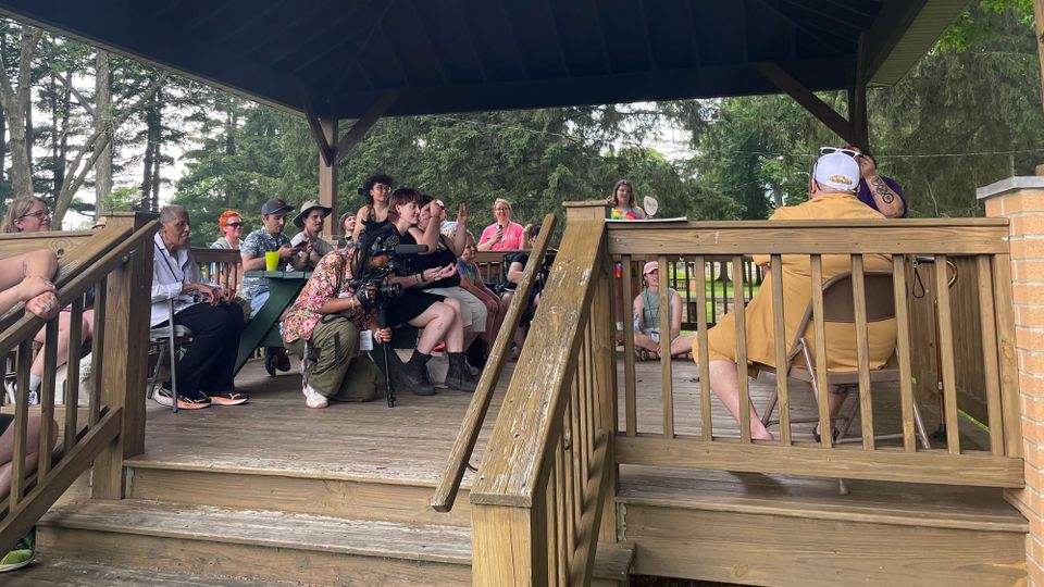 A wooden pavilion in a park on a summer day, with about a dozen people seated at picnic tables, watching a speaker