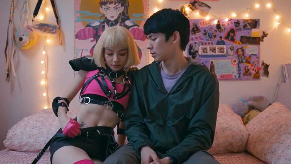 A young woman in pink and black punk gear and a young man in nondescript clothes, in her pink bedroom
