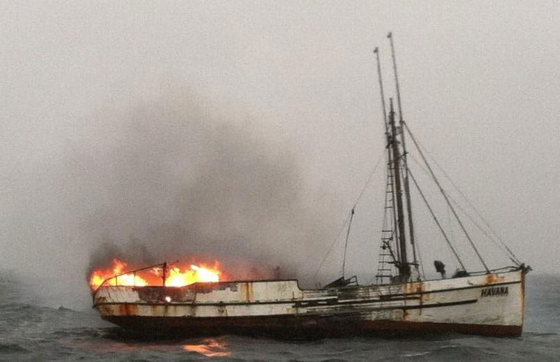 A fishing boat on the water, on fire, under a cloudy sky
