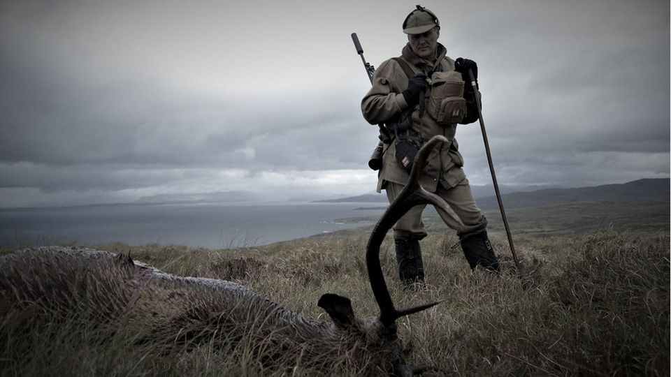 A deerstalker fully equipped for the hunt stands over the body of a buck, in a field by the sea under a cloudy sky