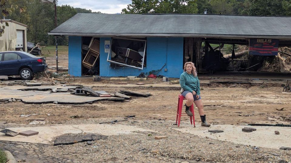 Root Bar proprietor Terri Dolan on a red barstool in front of the remains of the Root Bar after Hurricane Helene