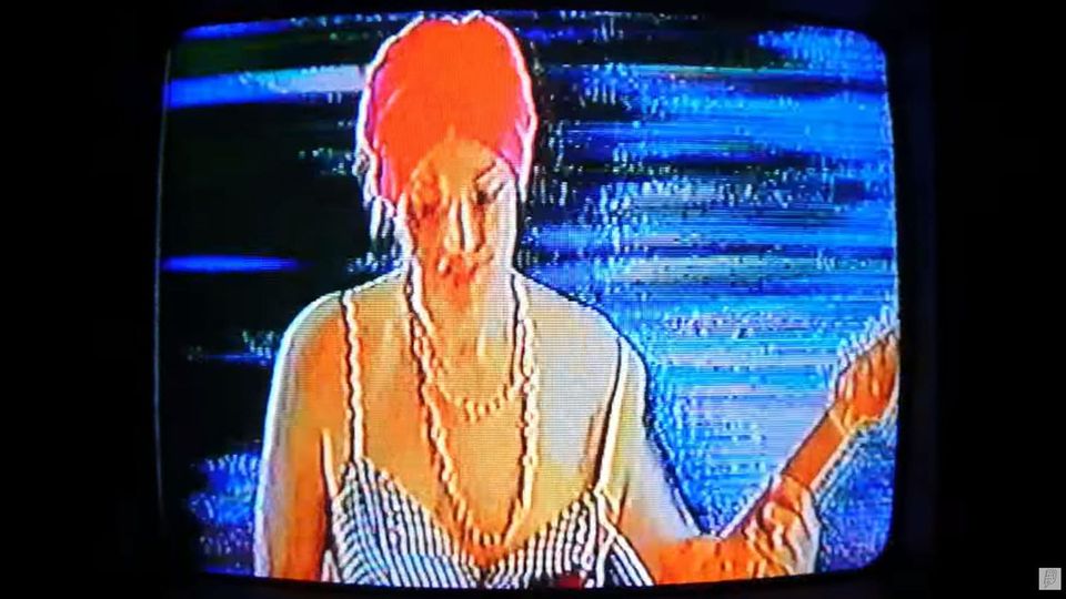 Grainy television image shows a guitarist in a red turban, gold chain and blue-and-white-striped spaghetti-strap dress
