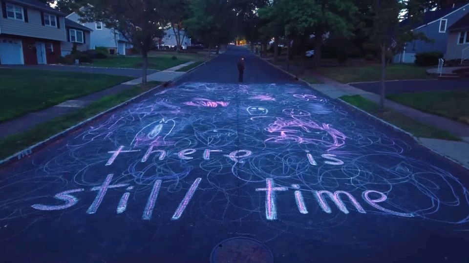 Scrawled sidewalk chalk on a tree-lined, twilit suburban street spells out 'there is still time'