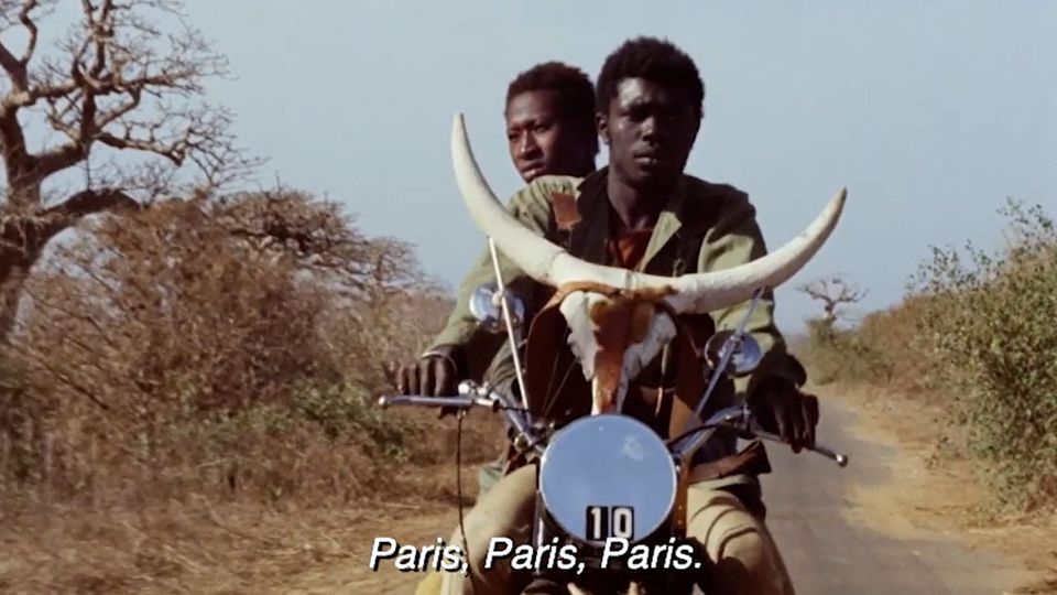 Two Senegalese men riding a motorcycle through sunlit country. The handlebars are decorated with bull horns.