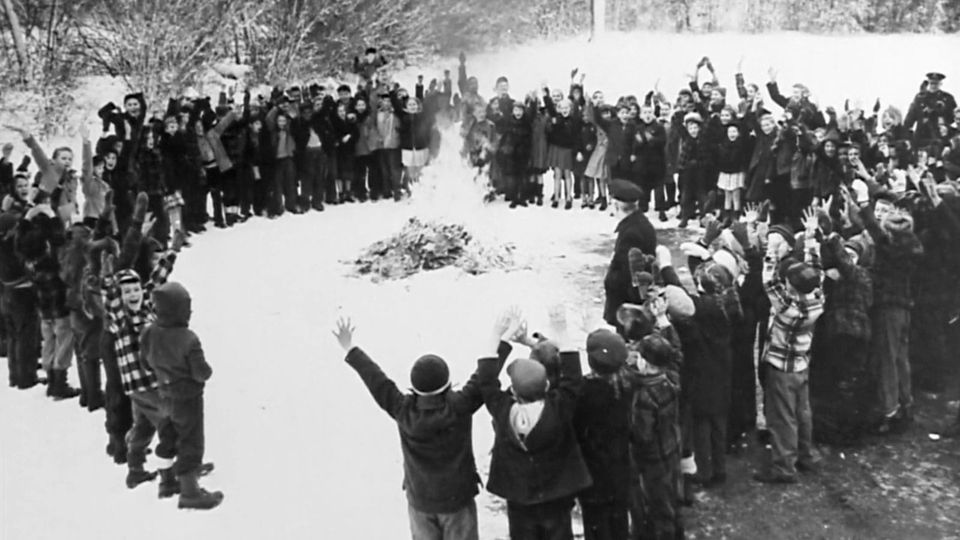 A black and white image of a circle of children cheering as a pile of comic books and magazines burn