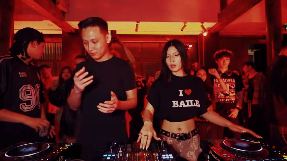 Two Chinese DJs dancing to their set in a red-light nightclub; a man in a black t-shirt and a woman in a black t-shirt reading 'I 💗 BAILE'