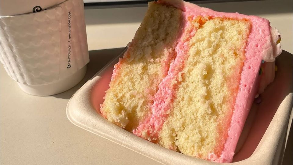 Takeout coffee cup beside a paper tray holding a large slice of golden layer cake with thick, pale bubblegum-pink frosting