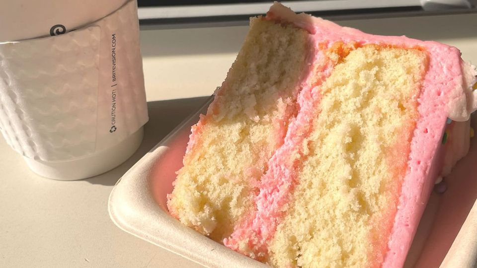 Takeout coffee cup beside a paper tray holding a large slice of golden layer cake with thick, pale bubblegum-pink frosting