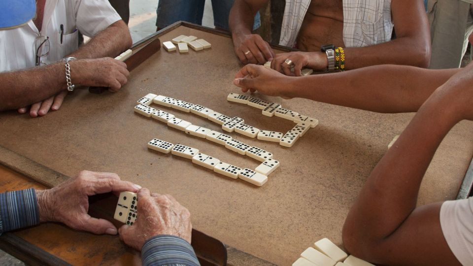 Four men playing dominoes at a small table in Havana, Cuba, in 2012; only their arms and hands are visible, some wearing watches, bracelets, and rings; the layout of the dominoes makes a "W"