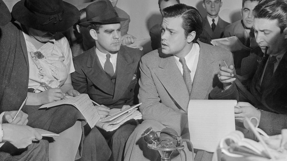 Black and white photo, 1938, of reporters crowded around Orson Welles, in a press conference to discuss the 'War of the Worlds' radio broadcast