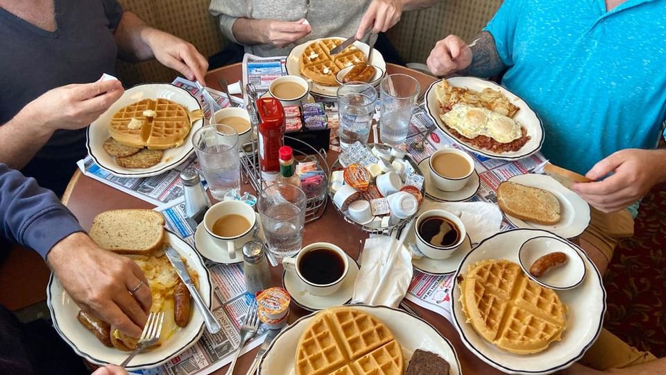 A table of breakfasters enjoying waffles, eggs, home fries, bacon, sausage, scrapple, toast and coffee
