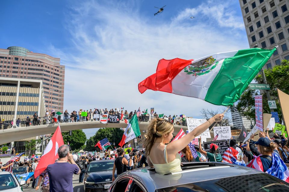 A woman emerging from the sunroof of a car and waving a Mexican flag on a gloriously sunny day in the crowd of the No Kings protest in L.A.