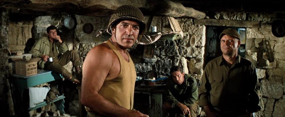 Telly in WWII Army helmet and undershirt, Rickles in Army cover and fatigues, and two other guys looking warily off camera 