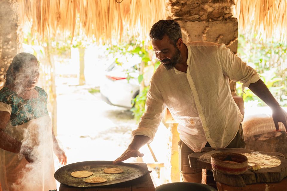 Author Michael Shaikh and a friend preparing cooking flatbreads on a wide pan in a semi-outdoor kitchen, sun streaming in behind them