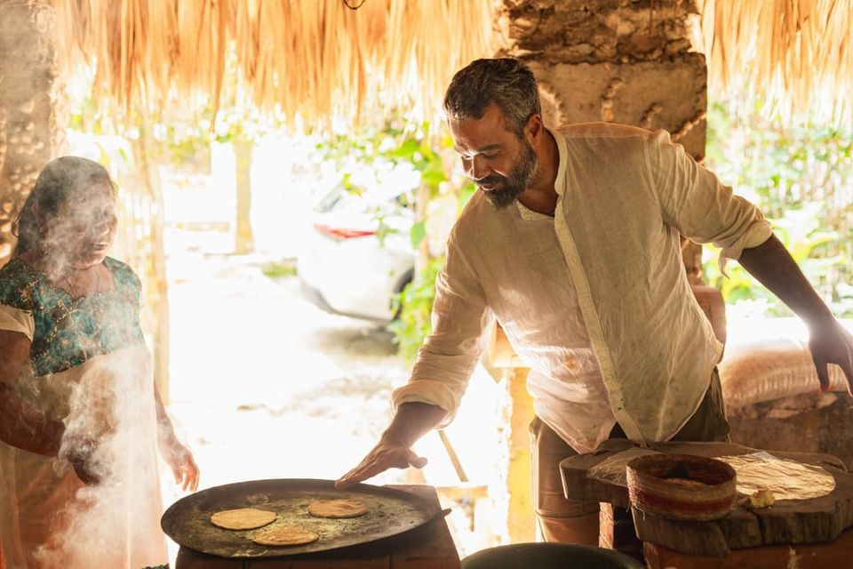Author Michael Shaikh and a friend preparing cooking flatbreads on a wide pan in a semi-outdoor kitchen, sun streaming in behind them
