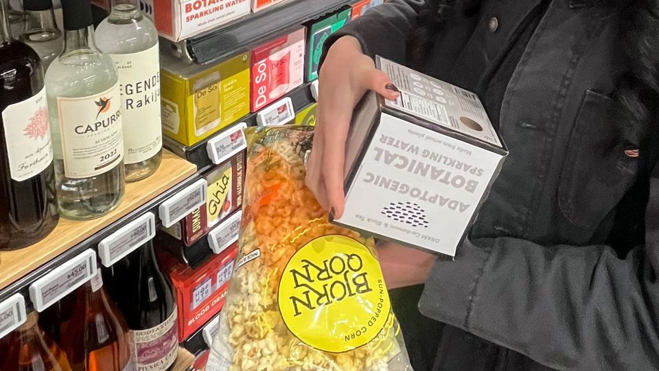 Colorful store shelves stuffed with pricey hooch and mixers; a shopper’s hands to the right holding a clear package of ‘Bjorn Qorn’ and a pack of Adaptogentic Botanical Sparkling Water’
