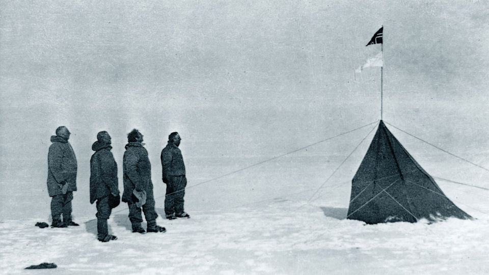 An icy South Pole scene from Roald Amundsen's 1911 expedition featuring ice, snow, a few rocks, and four men looking on at the Norwegian flag, aloft on a flagpole secured by long wires