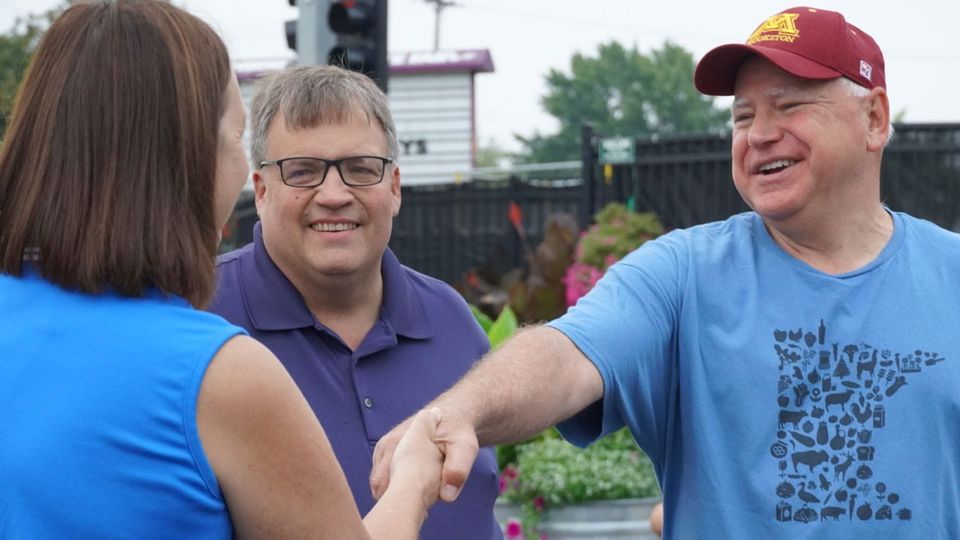 Tim Walz wearing a blue Minnesota t-shirt and red ball cap, and shaking hands with a woman in a blue shirt at the Minnesota State Fair, August 2023 