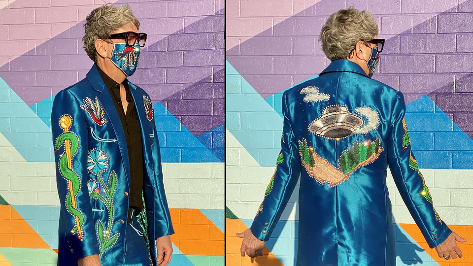 A man in thick black glasses and a suit and mask to match in violent electric blue embroidered with a spacecraft and a lurid green and gold caduceus