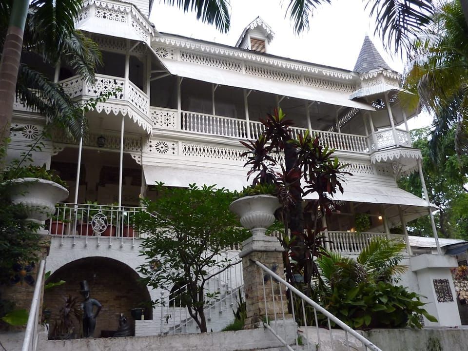 Grand wooden structure painted white, with white shade awnings, much gingerbread, set amid tropical greenery 