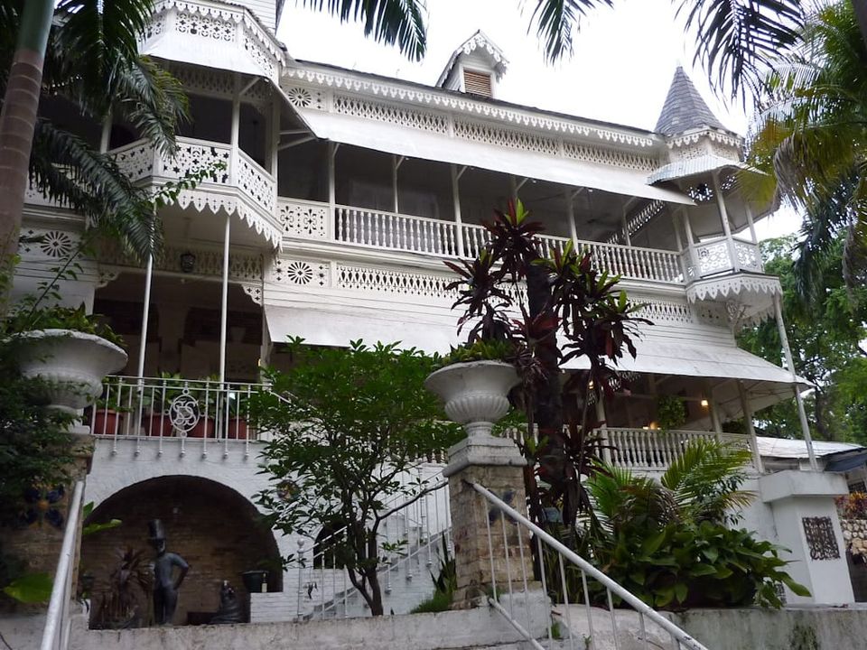 Grand wooden structure painted white, with white shade awnings, much gingerbread, set amid tropical greenery 