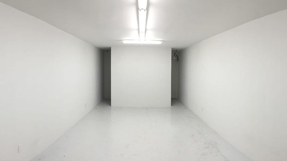 An empty gallery space, walls, floor and ceiling painted white. Fluorescent lighting glows in a line from back toward the viewer 