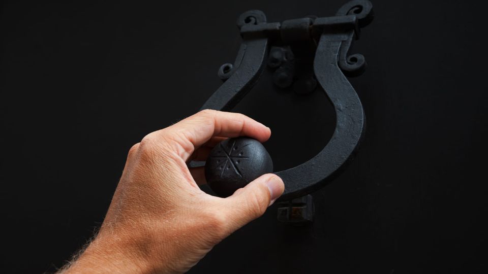 A hand lifting the rounded handle of a heavy cast iron knocker, about to strike a door